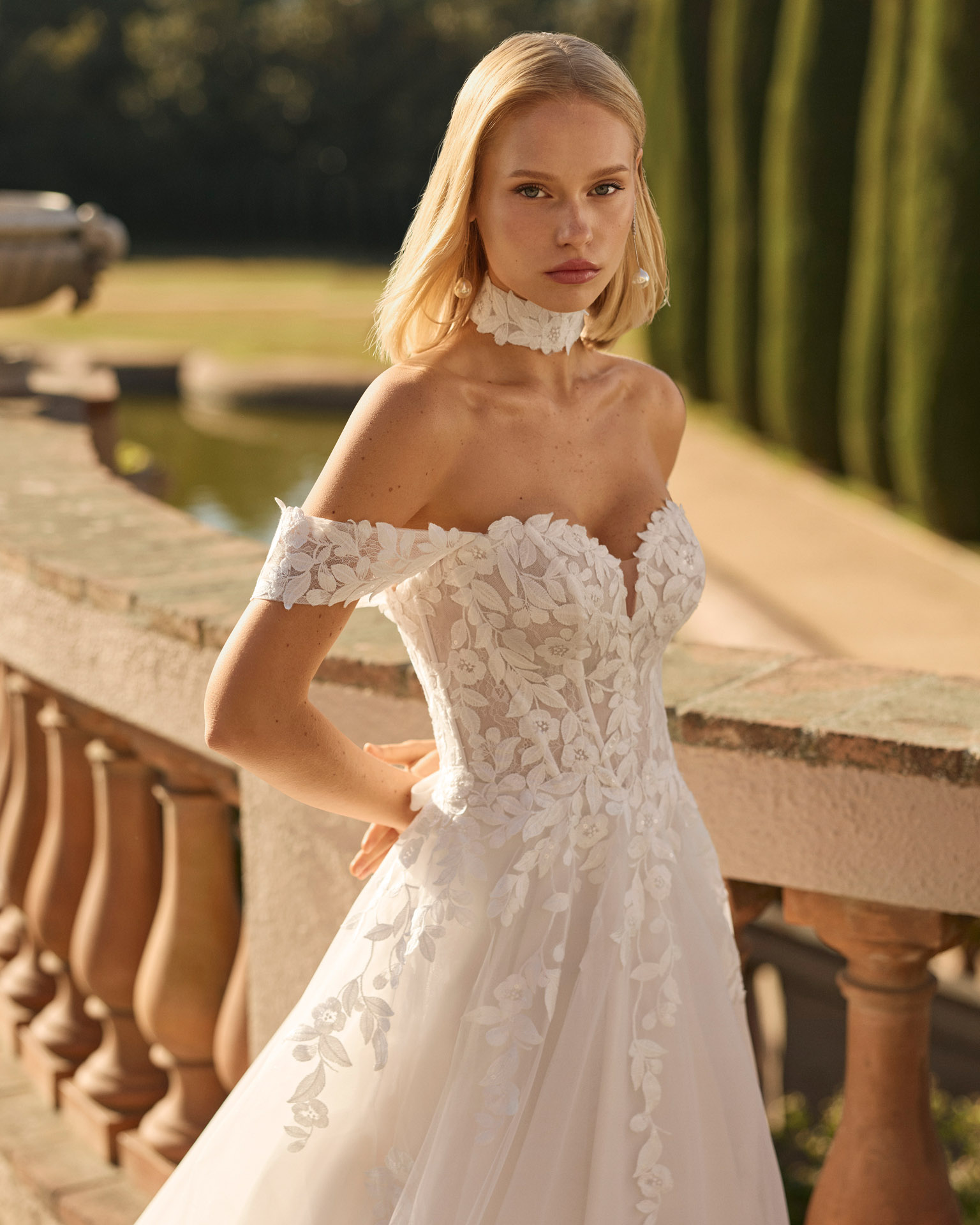 Vestido de novia Fresa de Luna Novias 2027 de corte A con escote barco, mangas caídas de encaje floral, delicado choker de encaje y falda de tul con aplicaciones florales.