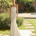 Vestido de novia Megan de Alma Novia 2027 con escote palabra de honor, silueta sirena y chaqueta de encaje desmontable de cuello alto y manga larga.