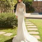 Vestido de novia Megan de Alma Novia 2027 con escote palabra de honor, silueta sirena y chaqueta de encaje desmontable de cuello alto y manga larga.
