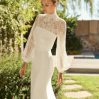 Vestido de novia Megan de Alma Novia 2027 con escote palabra de honor, silueta sirena y chaqueta de encaje desmontable de cuello alto y manga larga.