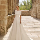 Vestido de novia Poppy de Adriana Alier 2027 con escote palabra de honor, cuerpo drapeado, falda de línea A y capa desmontable sobre los hombros.