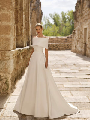Vestido de novia Poppy de Adriana Alier 2027 con escote palabra de honor, cuerpo drapeado, falda de línea A y capa desmontable sobre los hombros.