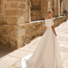 Vestido de novia Poppy de Adriana Alier 2027 con escote palabra de honor, cuerpo drapeado, falda de línea A y capa desmontable sobre los hombros.