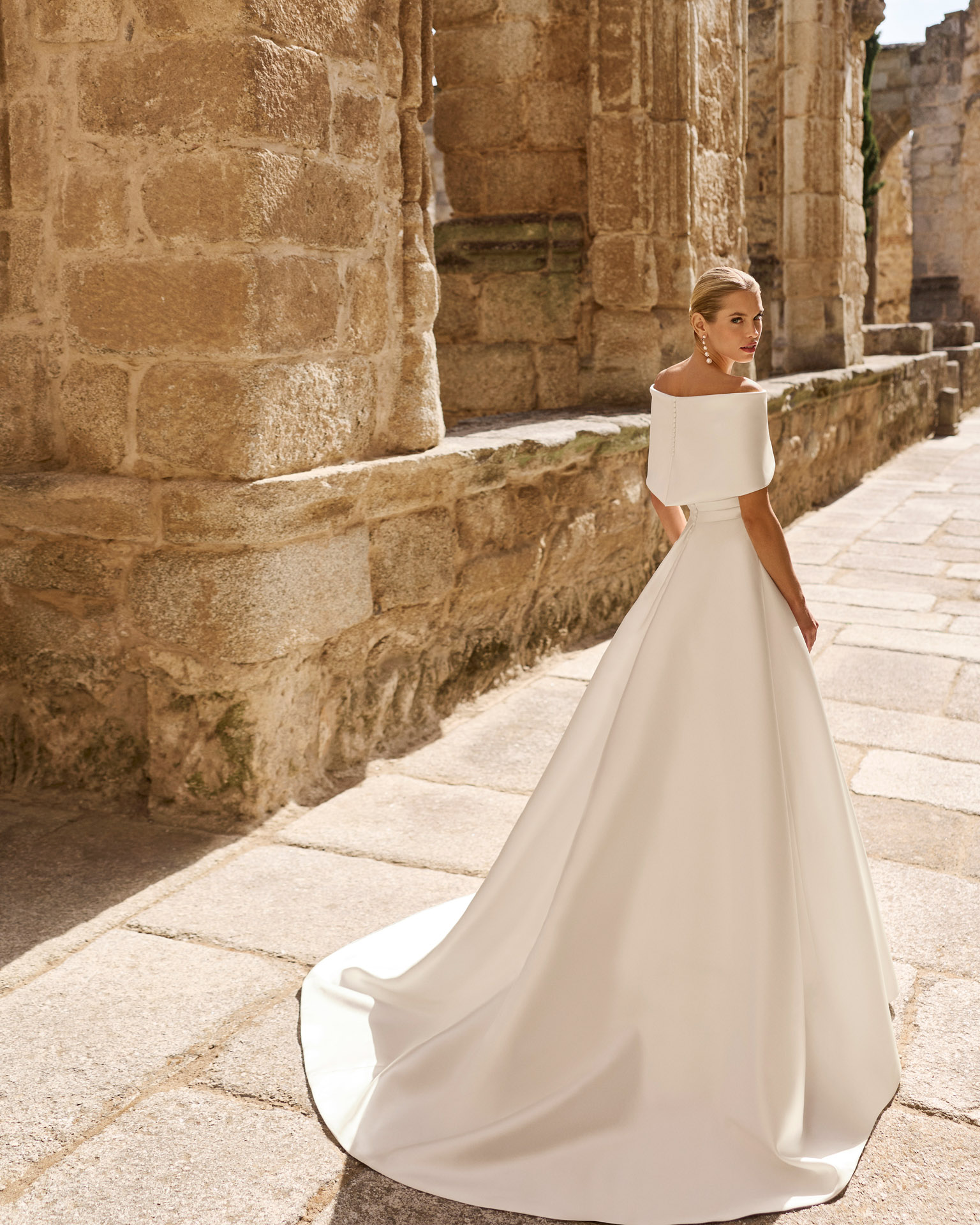 Vestido de novia Poppy de Adriana Alier 2027 con escote palabra de honor, cuerpo drapeado, falda de línea A y capa desmontable sobre los hombros.