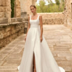 Vestido de novia Porto de Adriana Alier 2027 de línea A con escote cuadrado, abertura frontal, cintura de encaje y sobrefalda desmontable.