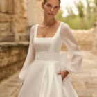 Vestido de novia Porto de Adriana Alier 2027 de línea A con escote cuadrado, abertura frontal, cintura de encaje y sobrefalda desmontable.