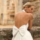 Vestido de novia Prado de Adriana Alier 2027 con cuello halter, espalda completamente descubierta, gran lazo posterior, falda de corte A con abertura lateral y tejido jacquard floral.