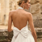 Vestido de novia Prado de Adriana Alier 2027 con cuello halter, espalda completamente descubierta, gran lazo posterior, falda de corte A con abertura lateral y tejido jacquard floral.