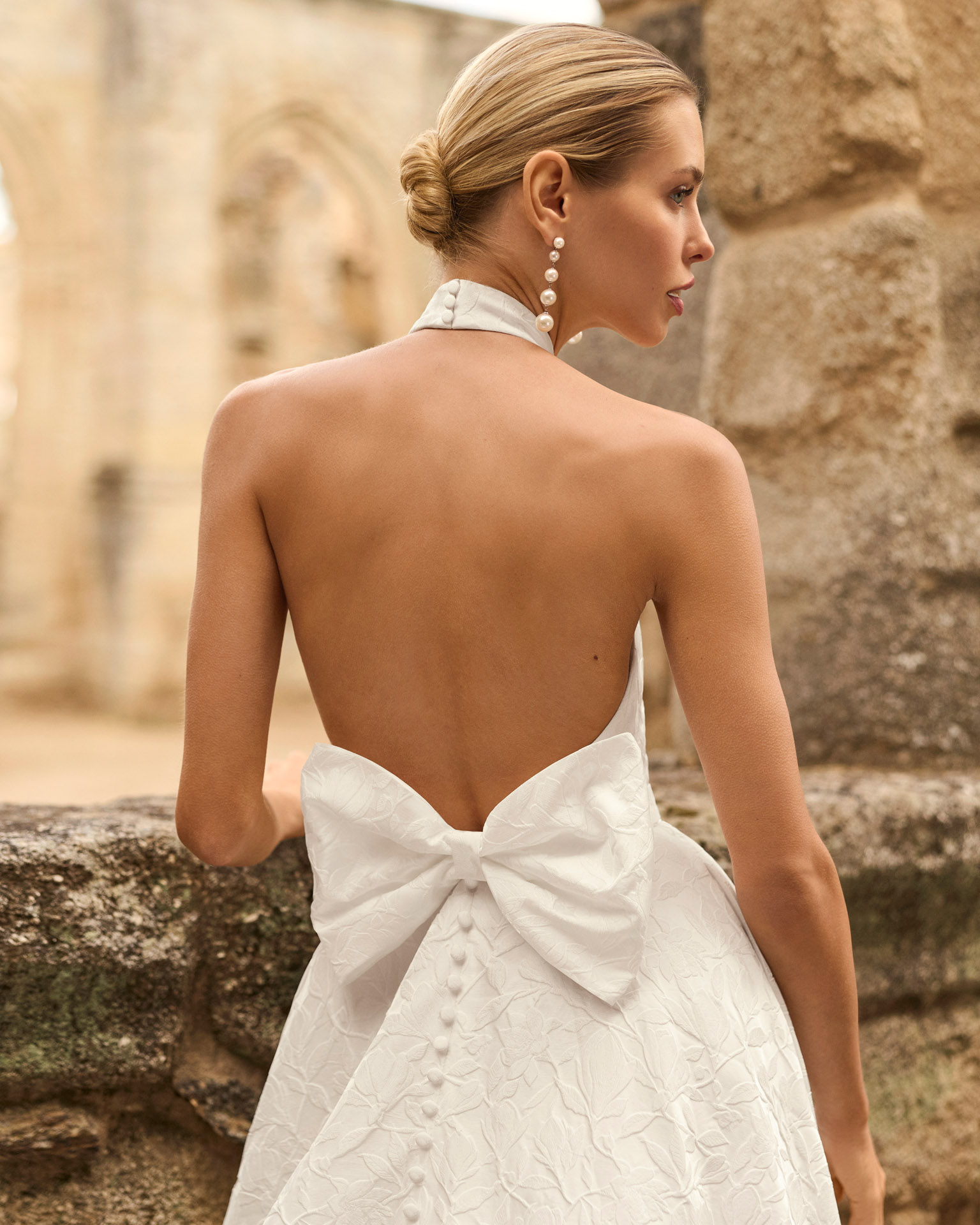 Vestido de novia Prado de Adriana Alier 2027 con cuello halter, espalda completamente descubierta, gran lazo posterior, falda de corte A con abertura lateral y tejido jacquard floral.