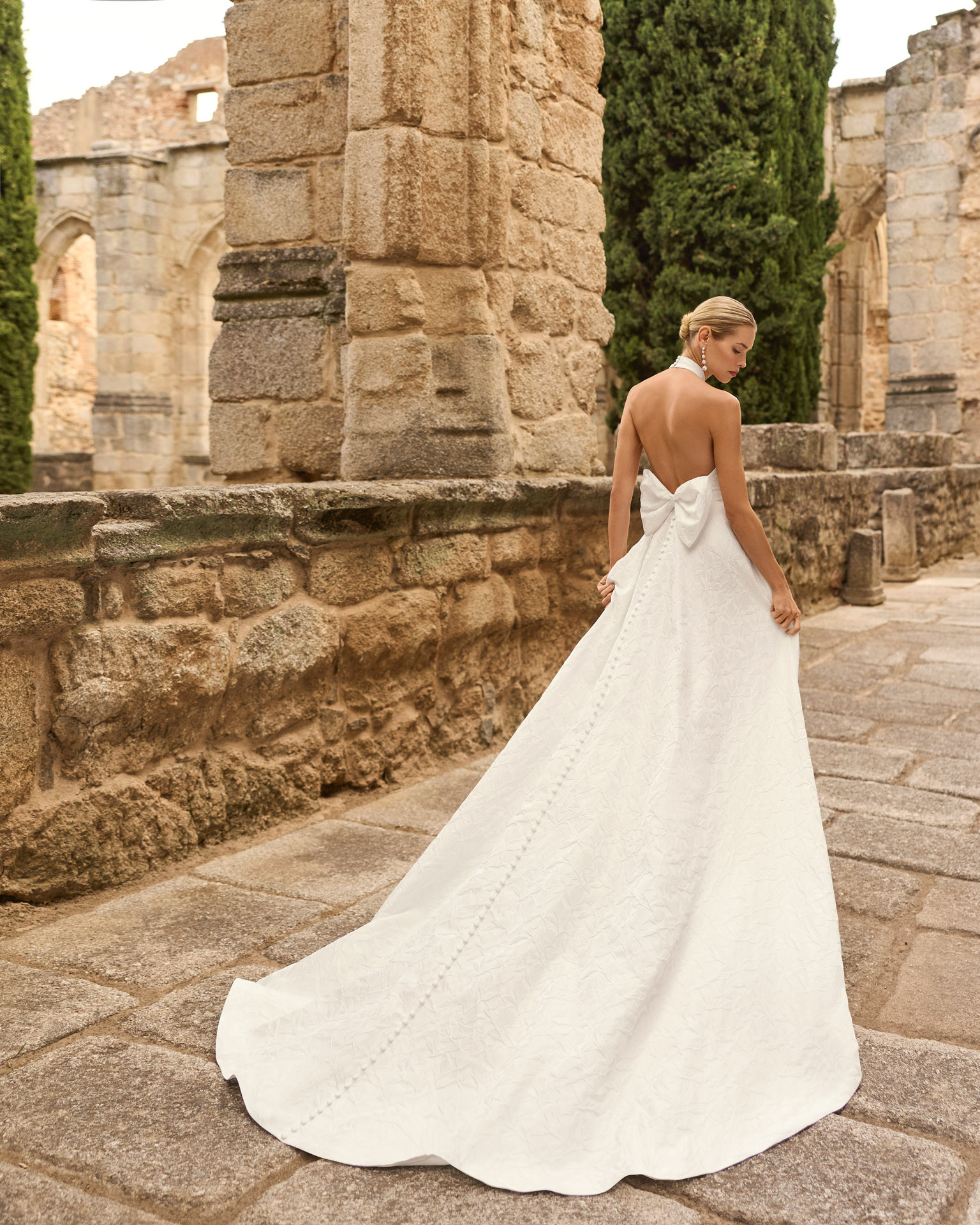 Vestido de novia Prado de Adriana Alier 2027 con cuello halter, espalda completamente descubierta, gran lazo posterior, falda de corte A con abertura lateral y tejido jacquard floral.