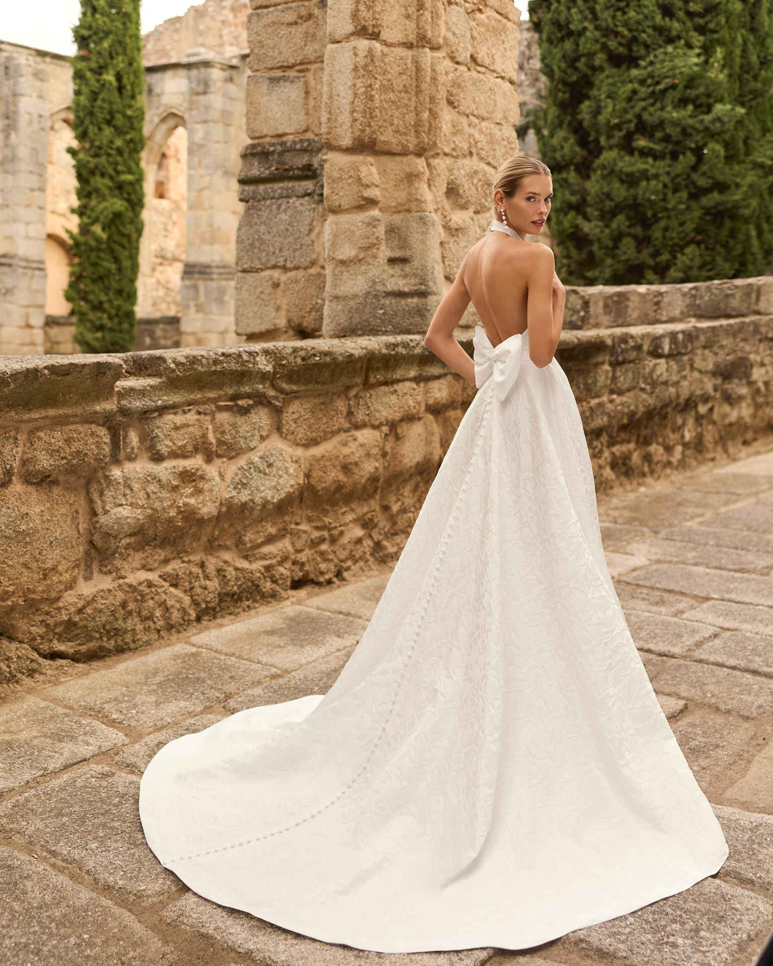 Vestido de novia Prado de Adriana Alier 2027 con cuello halter, espalda completamente descubierta, gran lazo posterior, falda de corte A con abertura lateral y tejido jacquard floral.
