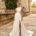 Vestido de novia Prado de Adriana Alier 2027 con cuello halter, espalda completamente descubierta, gran lazo posterior, falda de corte A con abertura lateral y tejido jacquard floral.