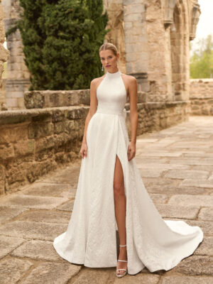 Vestido de novia Prado de Adriana Alier 2027 con cuello halter, espalda completamente descubierta, gran lazo posterior, falda de corte A con abertura lateral y tejido jacquard floral.
