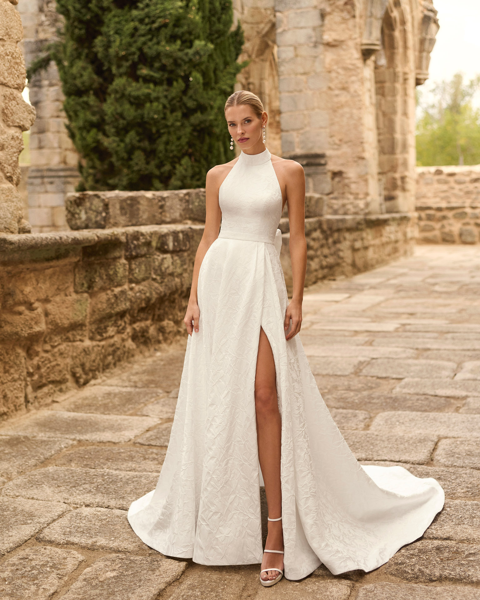 prado-vestido-novia-cuello-halter-espalda-descubierta-lazo-adriana-alier-2027.frente Vestido de novia Prado de Adriana Alier 2027 con cuello halter, espalda completamente descubierta, gran lazo posterior, falda de corte A con abertura lateral y tejido jacquard floral.