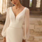 Vestido de novia Princes de Adriana Alier 2027 en crepé con escote en V, mangas largas de gasa, espalda en V con gran lazo y falda de línea A.