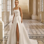 Vestido de novia Aire Barcelona 2027 de mikado con escote cuadrado, tirantes anchos, falda línea A con abertura delantera y detalle floral en la cintura.2